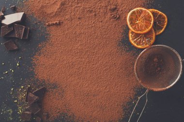 Kakao tozu koyu bir yüzeyi çikolata ve kuru portakal dilimleriyle kaplıyor. Bir elek kenarda oturur ve tatlı ya da içecek hazırlığı önerir..