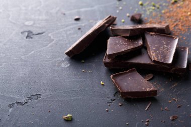 Kırık siyah çikolata parçaları siyah bir mutfak tezgahına saçılmış. Yakınlarda kakao tozu var, çikolatanın zengin renklerini vurguluyor..