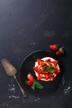 Yuvarlak bir tatlı siyah bir tabağa konur ve üzerine taze çilek konur. Tabağın yanında servis aleti var. Arka planda desenli bir yüzey var. Bazı çilekler etrafa saçılmış..