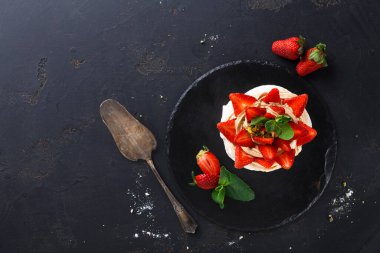 Yuvarlak bir tatlı taze çilek ve nane yapraklarıyla kaplıdır. Tatlının yanına eski bir servis bıçağı yerleştirilir. Arka planda birkaç dağınık çilek olan koyu bir yüzey var..