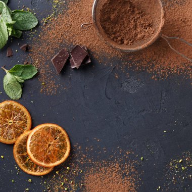 Nane yaprakları, kurutulmuş portakal dilimleri ve siyah bir mutfak masasında siyah çikolata parçaları düzenlenir. Kakao tozu etrafa saçılmış, aktif bir yemek pişirme sahnesi var..