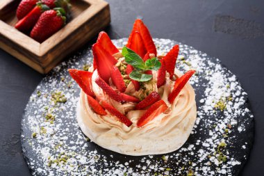 Bir tatlı siyah bir tabağa konur. Krem şanti içeriyor ve üzerinde taze çilek var. Tahta tepside ilave çilek ve yeşil bir garnitür bulunur..