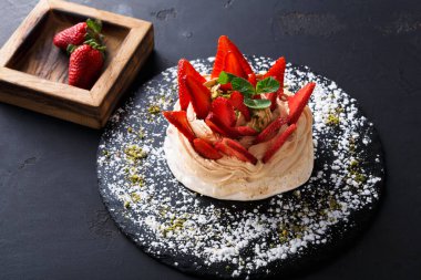 Yuvarlak bir tatlı, üzerinde dilimlenmiş çilekler ve bir tutam yeşillik olan siyah bir tabakta oturur. Yakınlardaki ahşap bir kutuda bütün çilekler var. Üstte nane var..