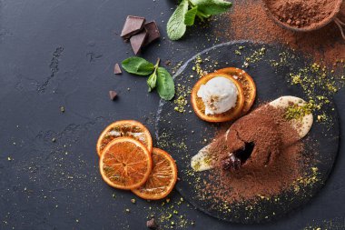 Tatlı tabağında çikolata tozu, bir kepçe dondurma ve kurutulmuş portakal dilimleri var. Nane yaprakları etrafa saçılmış. Ayar karanlık bir yüzeyde, yiyecek öğelerine zıtlık ekliyor.