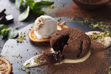 Çikolatalı bir tatlı siyah bir tabakta oturur. Tatlının ortasında erimiş bir merkez var ve yanında dondurma ve portakal dilimleri var. Yeşil nane yaprakları yemeğe renk katar.