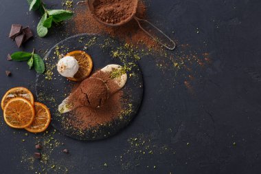 Koyu renkli bir yüzey, içinde dondurma, çikolata ve baharatlar bulunan bir tabak sunar. Portakal ve nane dilimleri renk katar. Sahnede detaylara dikkat eden bir tatlı sergileniyor..