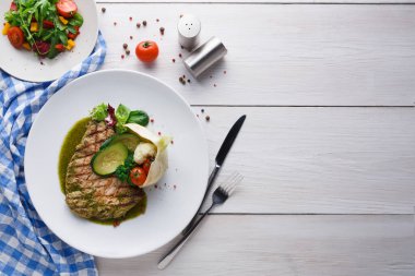 Bir tabak ızgara et taze salata ve sebzelerin yanında oturur. Ahşap bir masa ile ortam parlak. Yakınlarda sos kapları ve kiraz domatesleri var..