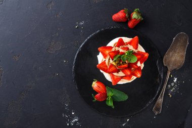 Yuvarlak katmanlı bir tatlı taze çilek dilimleri ve nane yapraklarıyla kaplıdır. Bütün çilekler tatlının yanına koyu bir yüzeye yerleştirilir. Gümüş servis kaşığı yakınlarda duruyor..