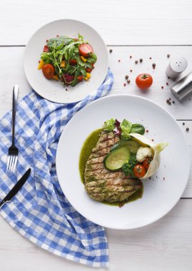 Izgara balık filetosu sebze ve sosla birlikte beyaz bir tabağa konur. Yanında küçük bir tabakta salata var. Masanın üzerinde mavi kareli bir peçete duruyor ve yanında biraz baharat var..