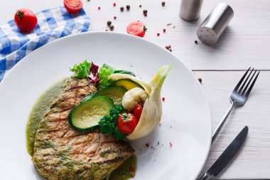 Beyaz tabakta servis edilen ızgara balığın olduğu bir tabak. Kabak ve domates gibi çeşitli sebzeler içerir. Dekorun yanında masa örtüsü ve baharatlar var..