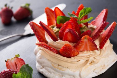 A dessert made of meringue topped with slices of fresh strawberries and mint leaves. Some whole strawberries are placed beside the dessert on a dark surface.