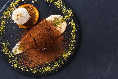 Tatlı tabağında yuvarlak bir çikolatanın yanında bir kepçe dondurma var. Tabak, dekorasyon için ezilmiş fındık ve portakal dilimleri. Sahne bir yemek deneyimini yakalar..