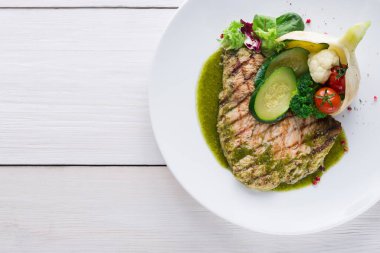 Izgara bir balık parçası beyaz bir tabağa konur. Tabakta salatalık, domates ve yeşillik gibi sebzeler yer alıyor. Yiyeceğin etrafına yeşil bir sos serpiştirildi. Ayarlar basit..