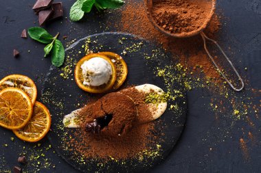 Sahnede siyah bir tabak çikolatalı tatlı, bir kepçe dondurma ve kurutulmuş portakal dilimleri gösteriliyor. Kakao tozu ve yeşil malzemeler etrafa serpilmiş. Parlak ışık dokuları vurgular.