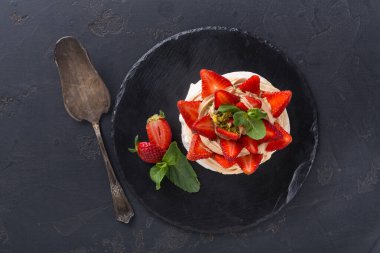 Yuvarlak bir tatlı taze çilek ile süslenmiş siyah bir tabakta oturur. Yakınlarda metal bir kaşık var. Yeşil nane yaprakları sunuma renk ekler.