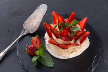 Tatlı, taze çileklerle çevrili krema katmanları içerir. Tabak, mutfaktaki siyah bir levhaya konur. Gümüş servis aparatı tatlının yanında oturur..