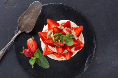 Krem şantili ve taze çilekli bir tatlı siyah bir tabakta sunulur. Nane yaprakları renk katar. Tabağın yanında gümüş servis aleti var..