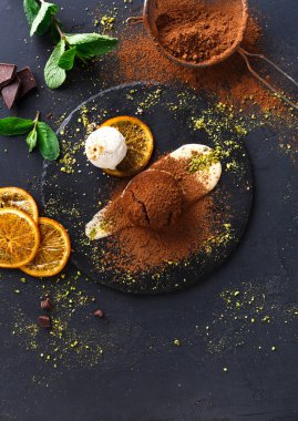 Yuvarlak siyah tabakta portakal dilimleri, muz ve üzerinde kakao tozu olan bir top dondurma var. Nane yaprakları ve çikolata parçaları kenarda görünür. Bu sahne bir mutfakta geçiyor..