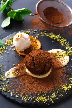 Bir tatlı, kakaoya bulanmış ve bir top dondurmayla servis edilmiş çikolatalı bir pasta içerir. Tabakta ezilmiş fındık ve restoran ortamında siyah bir tabakta bir portakal dilimi var..