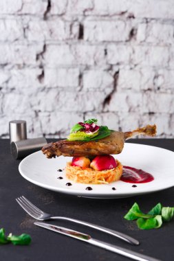 Bu tabakta soslu bir meyve yatağında ördek bacağı servis ediliyor. Arka plan dekora modern bir dokunuş katan desenli bir duvarı gösteriyor. Çatal ve bıçak tabağın yanında..