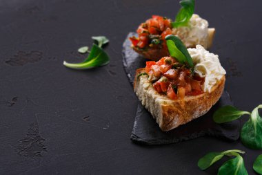 İki parça bruschetta koyu servis tabağına konur. Birinde otlu domates var. Diğerinde kremsi bir kaplama var. Tabağın etrafına taze yeşiller konur..
