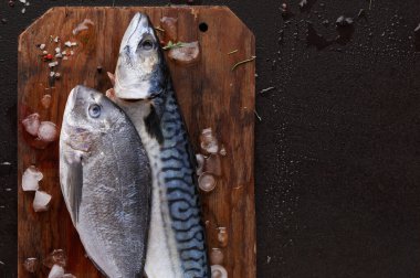 Tahta kesme tahtasının üzerinde yatan iki tür balık. Etrafları küçük buz parçaları ve biraz baharatla çevrili. Bu sahne mutfak veya pazar ortamında yemek hazırlıklarını gösteriyor..