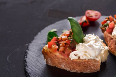 Koyu bir tabakta taze domates, fesleğen ve kremalı peynir servis edilir. Çeri domatesleri yakınlarda. Bu yemek zamanı başlangıç olarak uygundur..