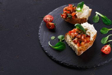 İki parça bruschetta siyah bir levhaya oturur. Birinde doğranmış domates ve bitkiler var, diğerinde ise kremalı dolgu. Çeri domatesleri ve yeşiller yakınlarda..