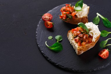 Hughchetta siyah bir servis tahtasına oturur. Etkinlikte üzerinde domates, ot ve peynir karışımı bir ekmek yer alıyor. Taze vişneli domates ve sebzeler yakınlarda..