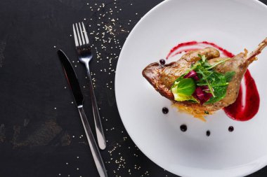 Izgara bir et parçası salatanın yanına beyaz bir tabağa konur. Tabak kırmızı sos ve siyah noktalarla süslenmiş. Gümüş çatal bıçak karanlık bir masada, tabağın yanında duruyor..