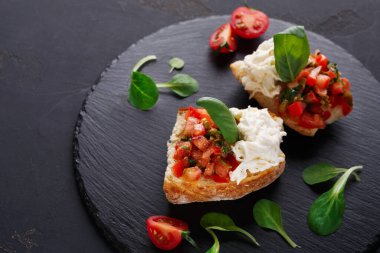 Siyah bir levhada iki parça bruschetta var. Birinde domates, ot ve yeşillik var. Diğerinde peynir ve taze malzemeler var. Çeri domatesleri yakınlarda..