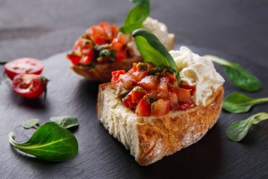 İki bruschetta parçası karanlık bir yüzeye yerleştirilir. Her birinde doğranmış domates ve şifalı otlar var. Krem peynir garnitür olarak eklenir. Taze yeşillikler ve kiraz domatesleri yakınlarda..