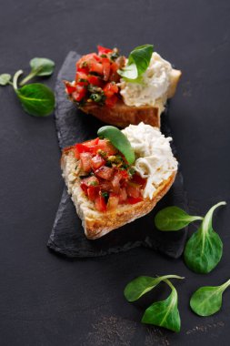 Koyu bir levhada iki parça bruschetta sergileniyor. Birinde doğranmış domates ve ottan bir tepsi, diğerinde ise kremalı bir sos var. Yeşiller tahtanın etrafına konur..