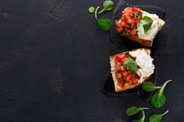 İki parça bruschetta koyu taştan bir tabağın üzerinde duruyor. Birinde doğranmış domates ve şifalı otlar var. Diğerinin kremsi beyaz bir tabakası var. Bruschetta 'yı çevreleyen yeşil yapraklar.