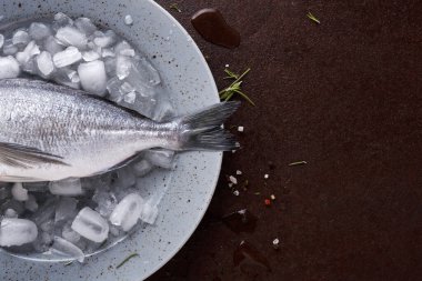Bir balık sığ bir kasede buzdan bir yatakta dinleniyor. Buz balığı taze tutmak için kullanılır. Tabağın etrafında az miktarda tuz ve otlar var. Mutfak ya da pazar gibi görünüyor..