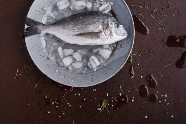 A whole fish rests on crushed ice inside a gray plate. The table is wooden with some spices and herbs scattered around. It is ready for cooking or meal preparation.