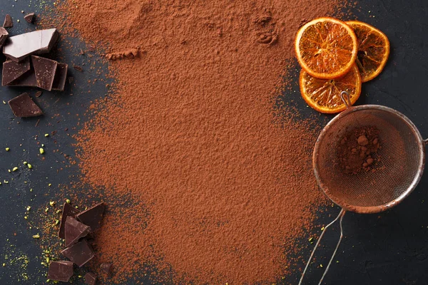 Siyah bir yüzeye yayılmış kakao tozu yakınında siyah çikolata parçaları düzenlenir. Kurutulmuş portakal dilimleri güzel bir elek ile yan yana oturur. Bu kurulum, pişirme veya tatlı malzemelerini gösterir.