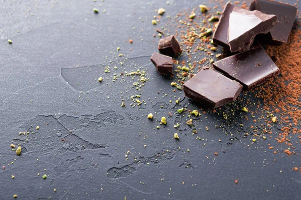 Parçalanmış siyah çikolata parçaları karanlık bir mutfak tezgahına saçılmış. Çikolatanın etrafında ezilmiş fındık ve kakao tozu var. Doğal ışık sahneyi aydınlatır..