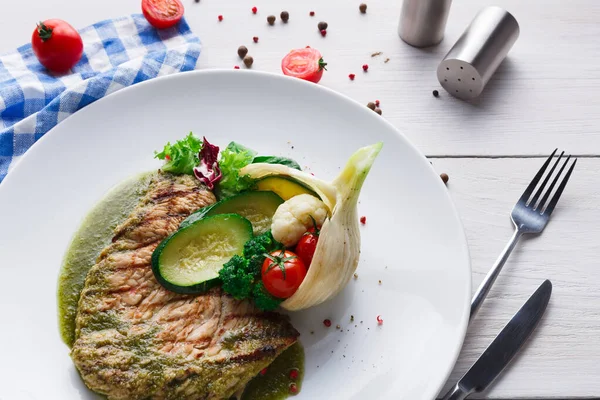 Beyaz tabakta servis edilen ızgara balığın olduğu bir tabak. Kabak ve domates gibi çeşitli sebzeler içerir. Dekorun yanında masa örtüsü ve baharatlar var..