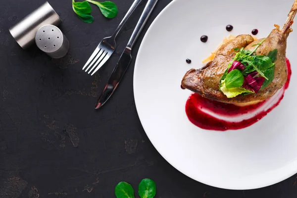 Güzel bir ördek tabağı beyaz bir tabakta oturur. Yemek sosu ve garnitür için taze yeşillikleri içeriyor. Sarımsak ezici ve çatal bıçak takımı karanlık bir masanın üzerindedir..