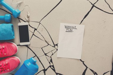 Antrenman planı, kırık bir zeminde spor malzemelerinin yanında bir kağıt üzerinde yapılır. Malzemeler arasında halterler, su şişesi ve spor ayakkabıları var. Kulaklık ve akıllı telefon da görülebilir..