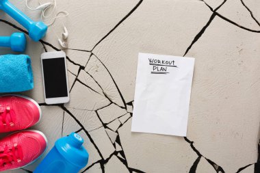 Spor salonu ekipmanlarıyla çevrili çatlak bir kata bir çalışma planı yerleştiriliyor. Yakınlarda bir çift pembe ayakkabı, mavi bir havlu, halterler, bir telefon ve bir su şişesi var..