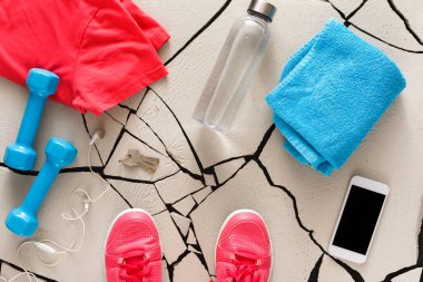Red workout shirt, blue towel, and light blue dumbbells are arranged on a cracked floor. A water bottle is nearby along with a phone and keys, ready for an exercise session.