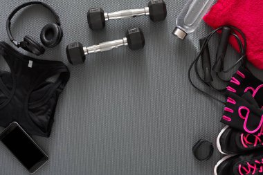 Items for workout are set on the gym floor. There are dumbbells, a water bottle, a towel, headphones, and fitness gear. A smartphone and accessories are also present, ready for use.