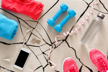 Bright workout clothes, weights, water bottle, phone, earphones, and keys are arranged on a cracked floor. This setup shows preparation for a workout session in a home setting.