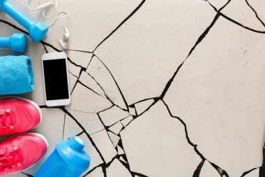 Bright pink sneakers sit next to blue dumbbells and a water bottle on a cracked floor. A towel and headphones are also nearby. A smartphone rests in the center of the setup.