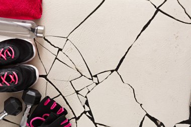 Bir sahne çatlak bir zeminde egzersiz malzemelerini gösteriyor. Pembe aksanlı siyah ayakkabılar, dambıllar, havlu ve su şişesi var. Bu kurulum bir çalışma ortamını temsil ediyor.