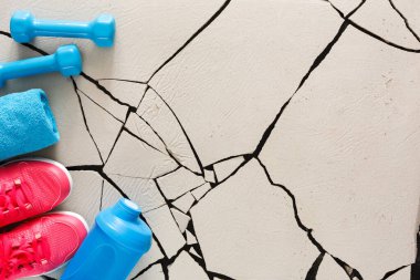Workout gear is laid out on a cracked floor. There are two blue dumbbells, a water bottle, a blue towel, and bright pink shoes. This setup is ready for an exercise session at the gym.