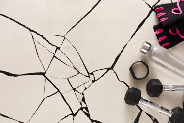 Bir çift dambıl, bir su şişesi ve spor eldivenleri görünür çatlaklarla bir spor salonuna yerleştirilir. Eşyalar yoğun bir egzersiz rutinine hazırlık gösteriyor..