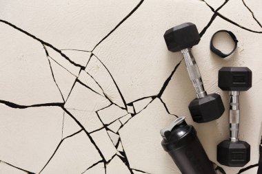 İki siyah dambıl ve bir su şişesi çatlamış beyaz bir yüzeyde dinleniyor. Ögeler fitness ve egzersiz odaklı bir egzersiz ayarını öneriyor.